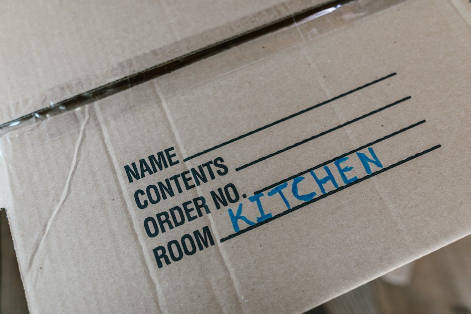 A close-up view of a cardboard box used in home relocation, showing a printed label with fields for 'Name,' 'Contents,' 'Order No,' and 'Room,' with the handwritten indication 'KITCHEN' in blue ink ne