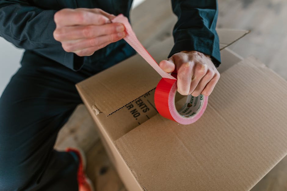 A close-up view of a cardboard box used in home relocation, showing a printed label with fields for 'Name,' 'Contents,' 'Order No,' and 'Room,' with the handwritten indication 'KITCHEN' in blue ink ne