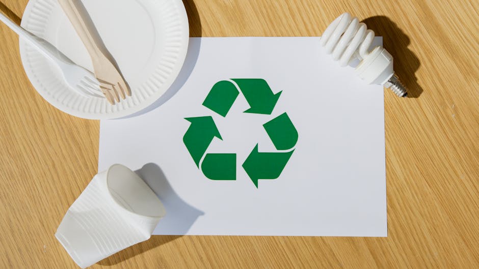 A flat lay image on a wooden surface showing a white sheet of paper with a large green recycling symbol printed on it, surrounded by various household waste items. In the top left corner, there is a w